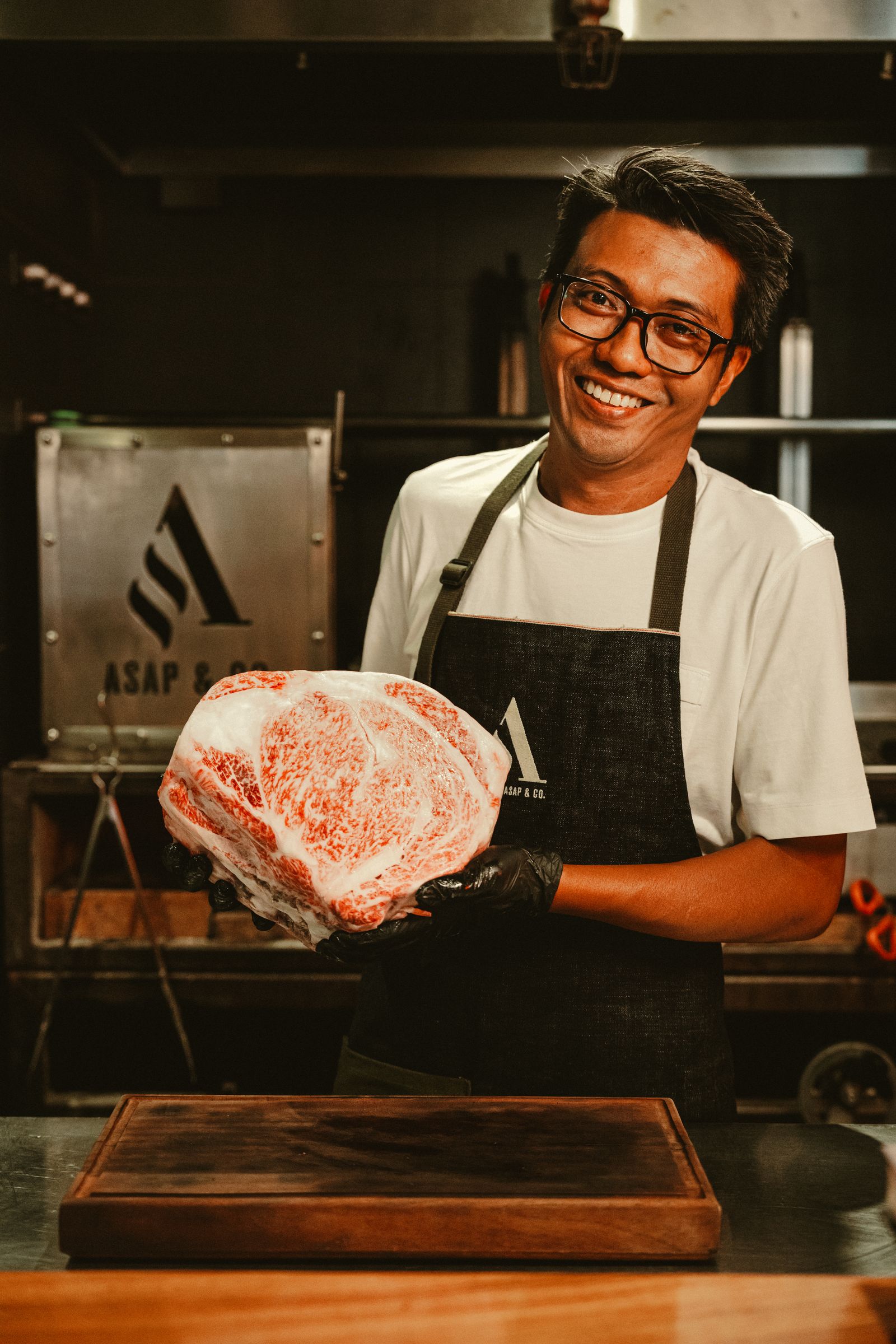 A chef in an ASAP & Co. apron presenting a marbled Wagyu cut at the grill